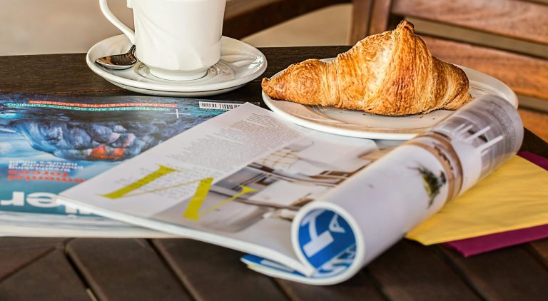 Enjoy a peaceful morning with coffee, croissant, and reading material on a wooden table.